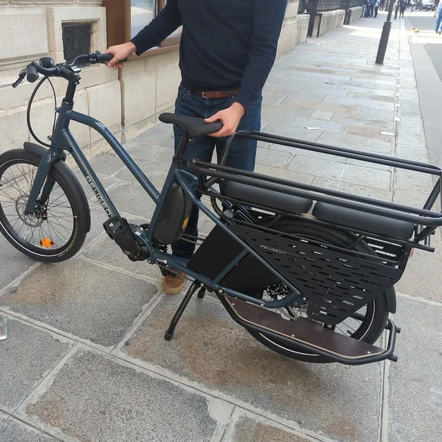 Un homme tient par le guidon et la selle un vélo longtail électrique gris sur un trottoir pavé